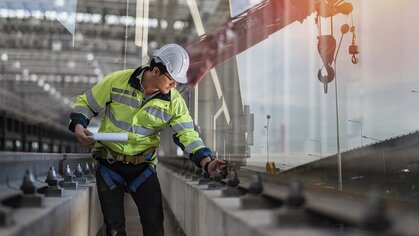 Photograph of a man wearing a white safety helmet and a yellow safety vest who inspects railroad tracks in an urban environment.